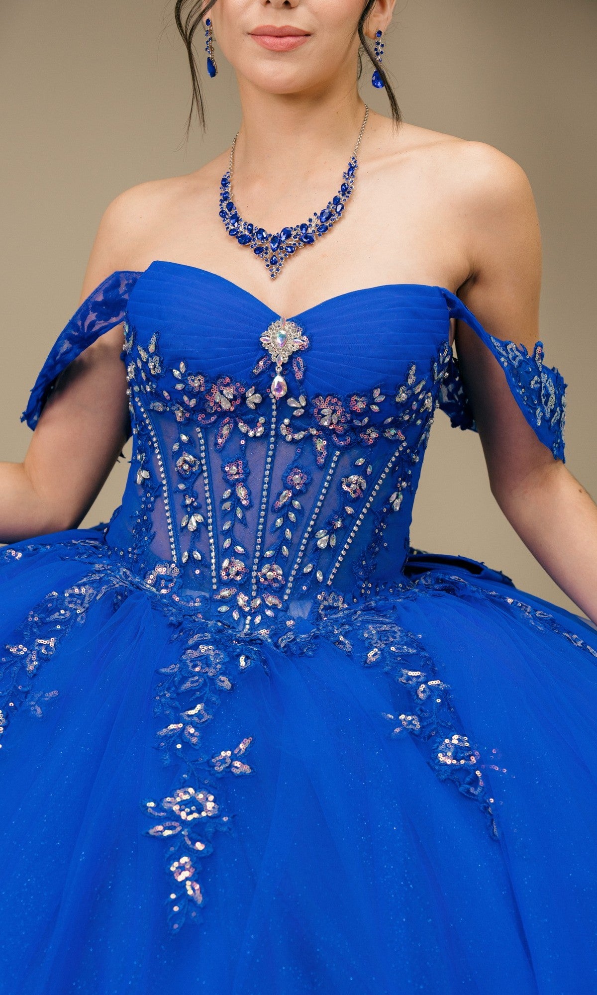 A woman wears the Quinceanera Dress 8405JQ By Cinderella Couture, a vibrant blue off-the-shoulder gown with beaded floral appliqué and matching blue jewelry—perfect for a quinceanera ball gown look.